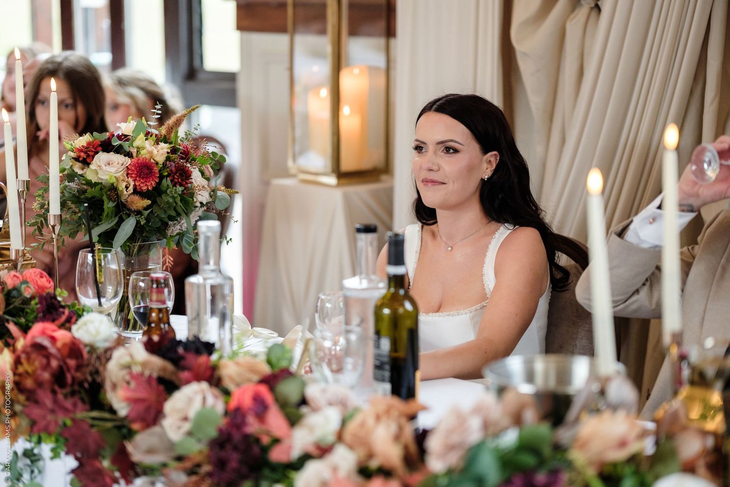 Bride in white dress seated at elegantly decorated wedding reception table with floral arrangements and candles.