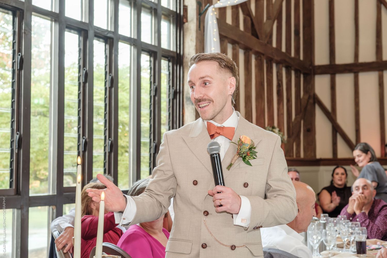 Groom in tan suit with orange bow tie gives speech holding microphone at elegant wedding reception venue.