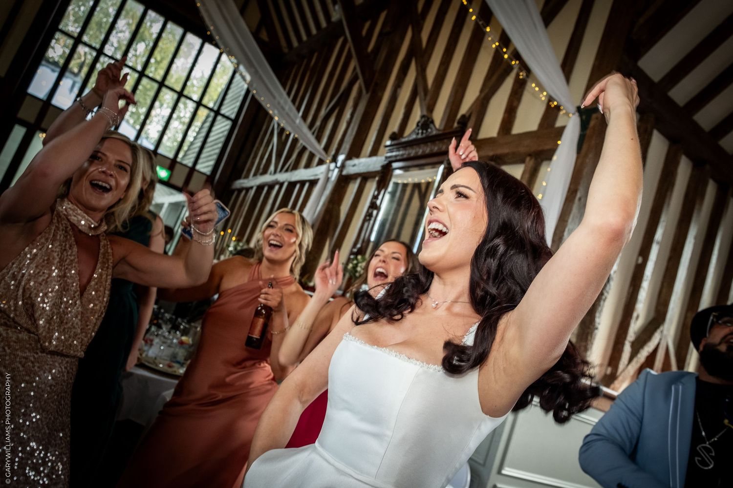 Bride in white dress celebrates joyfully with bridesmaids at wedding reception in a rustic timber-framed venue.