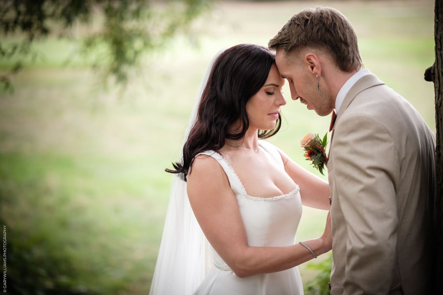 A bride and groom share an intimate moment, touching foreheads, dressed in white gown and tan suit outdoors.