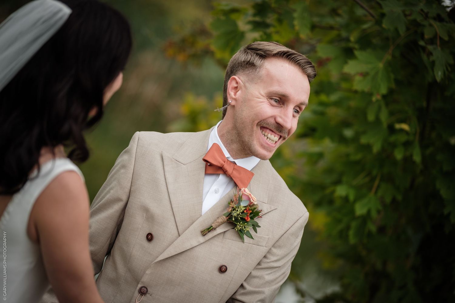 Smiling groom in beige double-breasted suit with coral bow tie and boutonniere standing with bride outdoors.