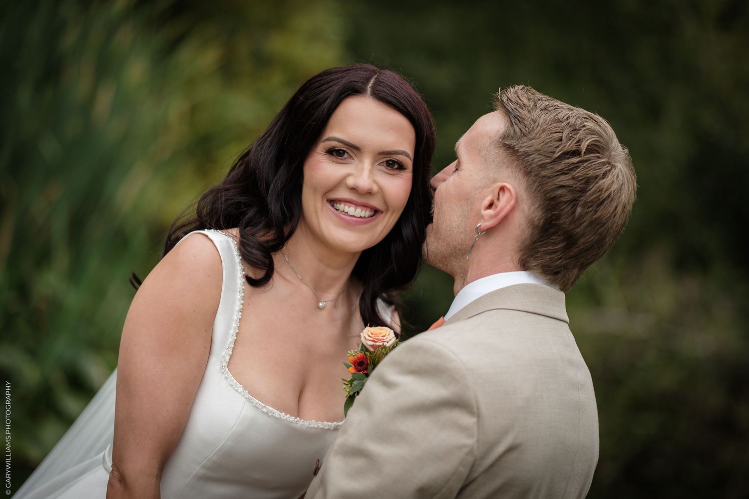 Groom in beige suit kissing bride's cheek as she smiles, outdoor wedding portrait with green foliage background.