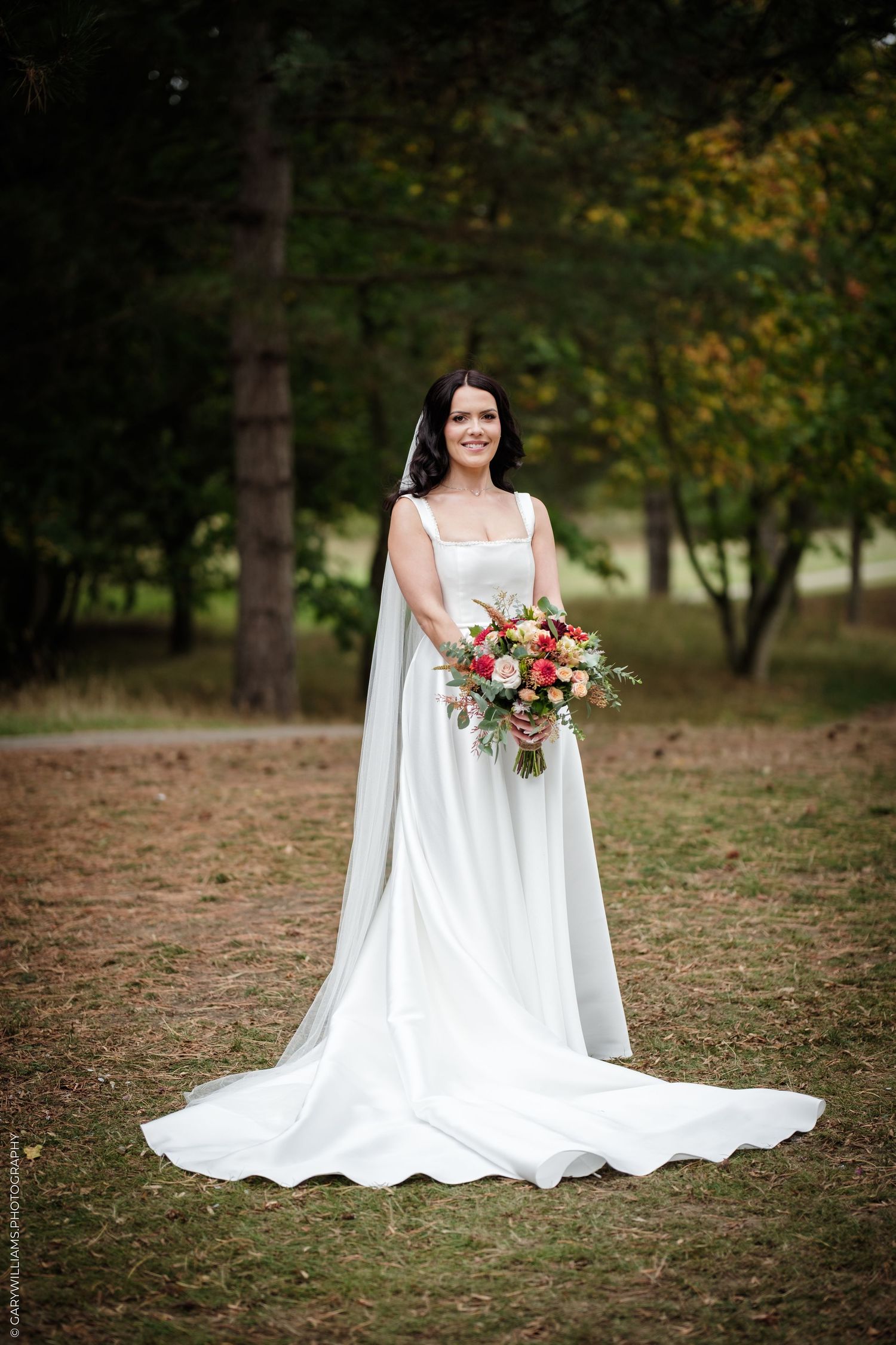 Bride in white gown holding colorful bouquet standing in a forest setting on her wedding day.