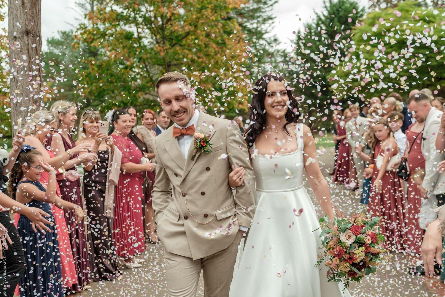 Bride and groom walk through confetti shower surrounded by smiling wedding guests outdoors.