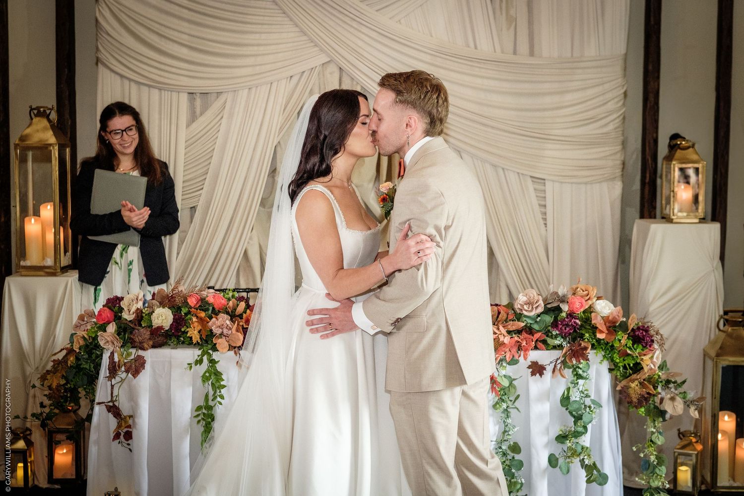 Bride and groom share first kiss at altar with floral arrangements, candles, and officiant in elegant draped venue.