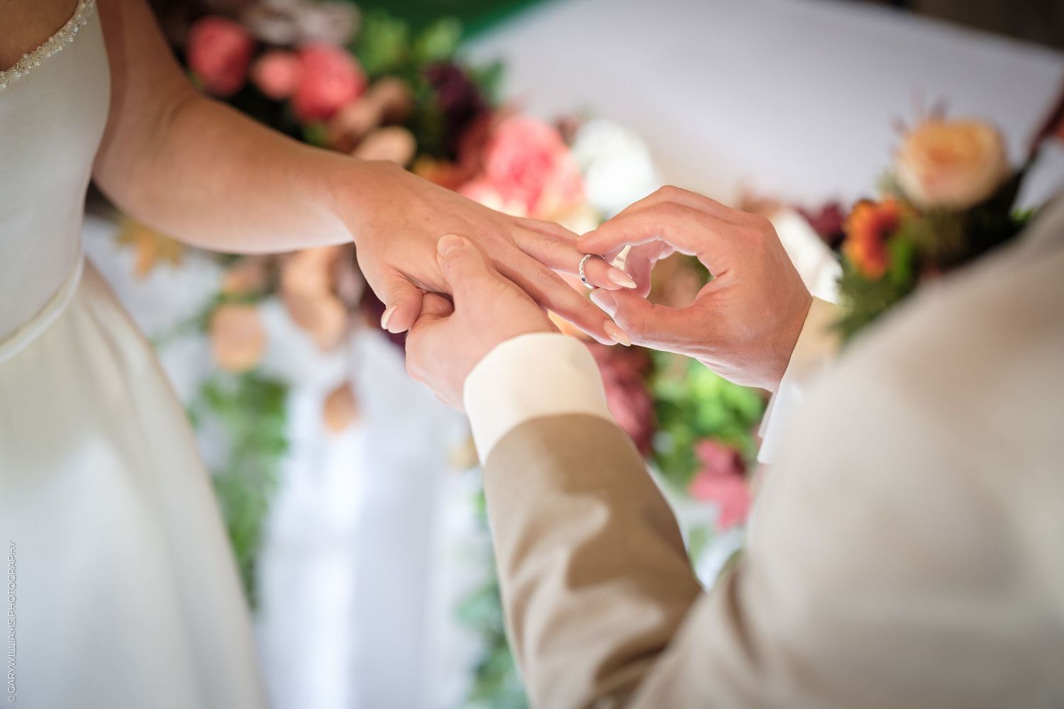 Bride placing a wedding ring on groom's finger during ceremony with floral backdrop.