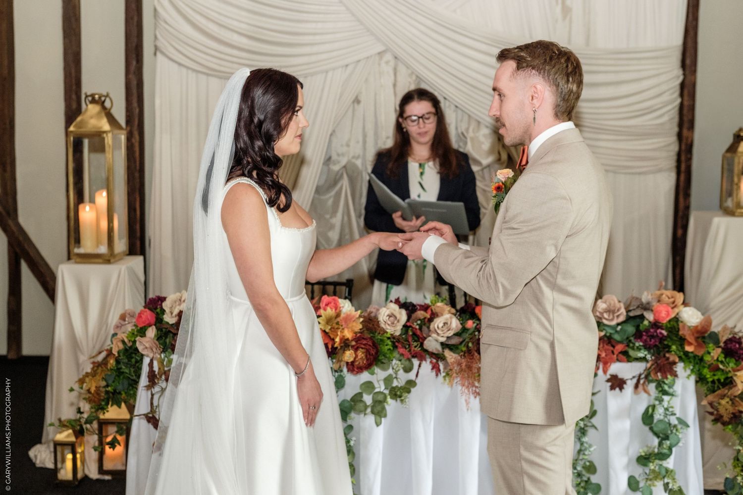 Bride and groom exchange rings at elegant wedding ceremony with floral arrangements and officiant in background.
