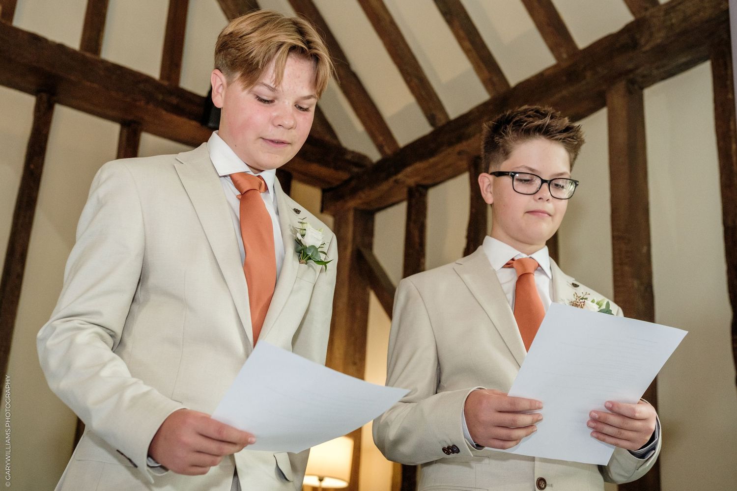 Two boys in matching beige suits and orange ties reading papers at a wedding ceremony.