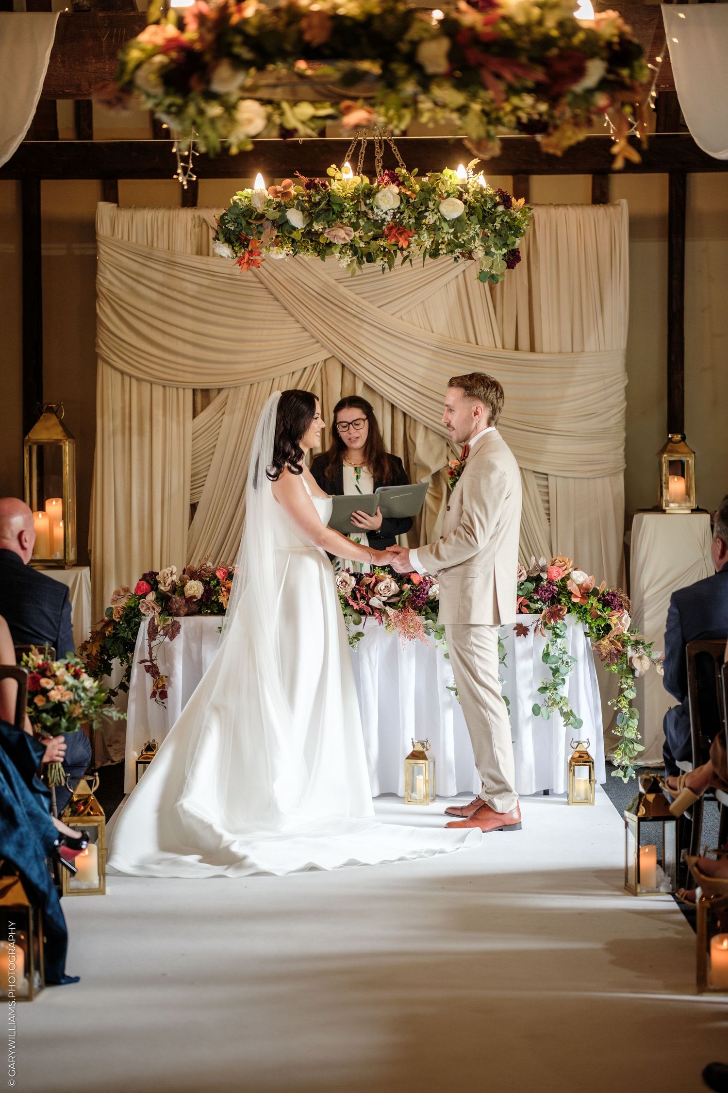 Bride and groom exchange vows at elegant indoor wedding ceremony with floral arch and draped fabric backdrop.