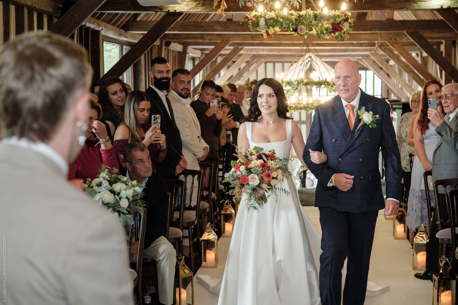 Bride walks down the aisle with her father in a rustic barn wedding ceremony, guests watching and smiling.