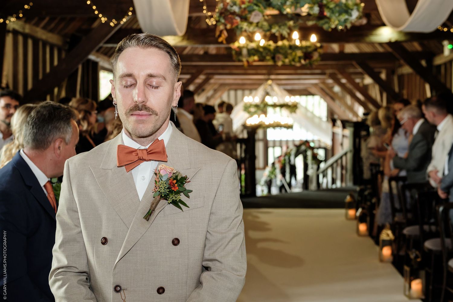 Groom in beige double-breasted suit with orange bow tie and boutonniere stands at wedding altar in rustic barn venue.