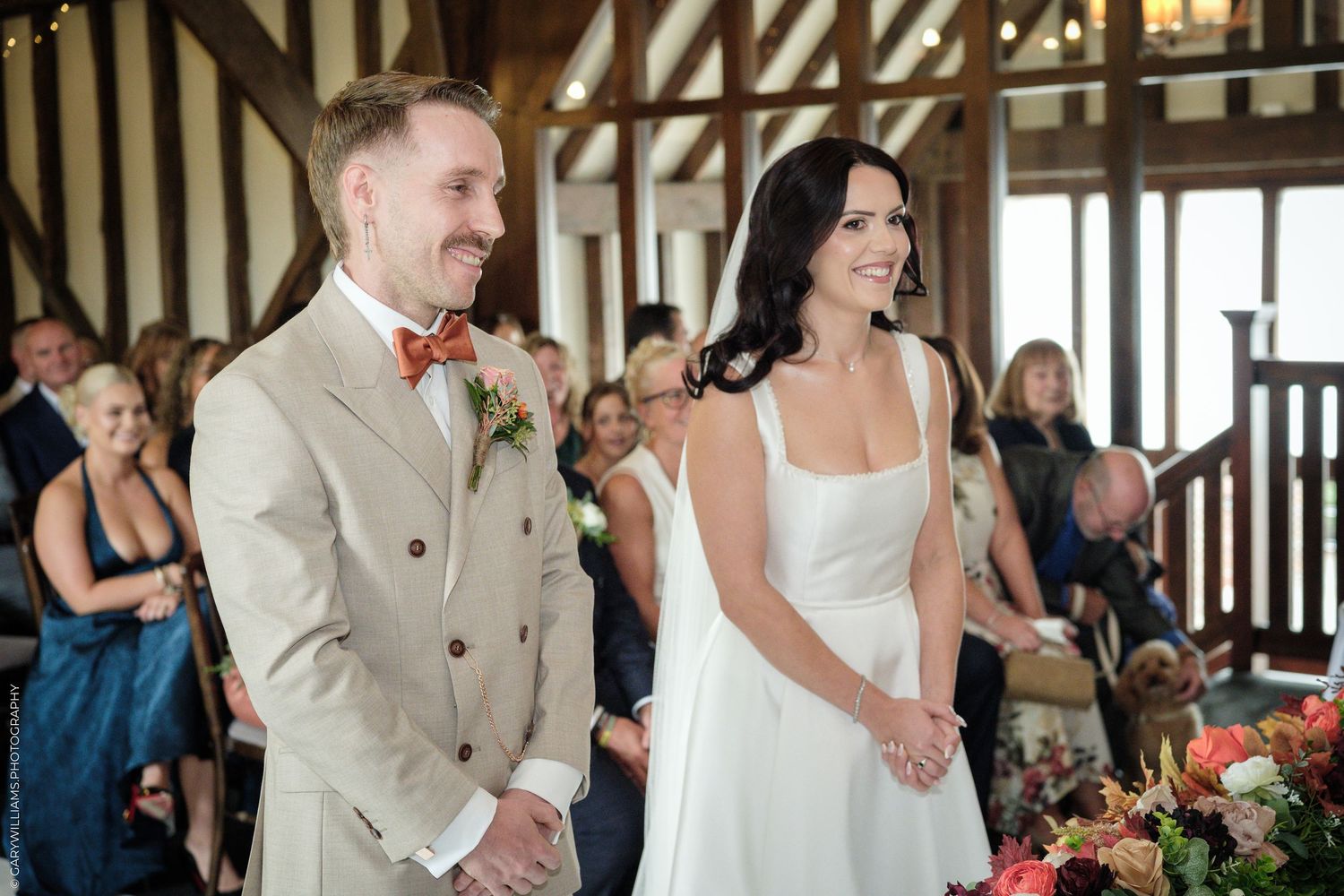 Bride and groom smiling during indoor wedding ceremony in rustic venue with floral arrangements and seated guests.