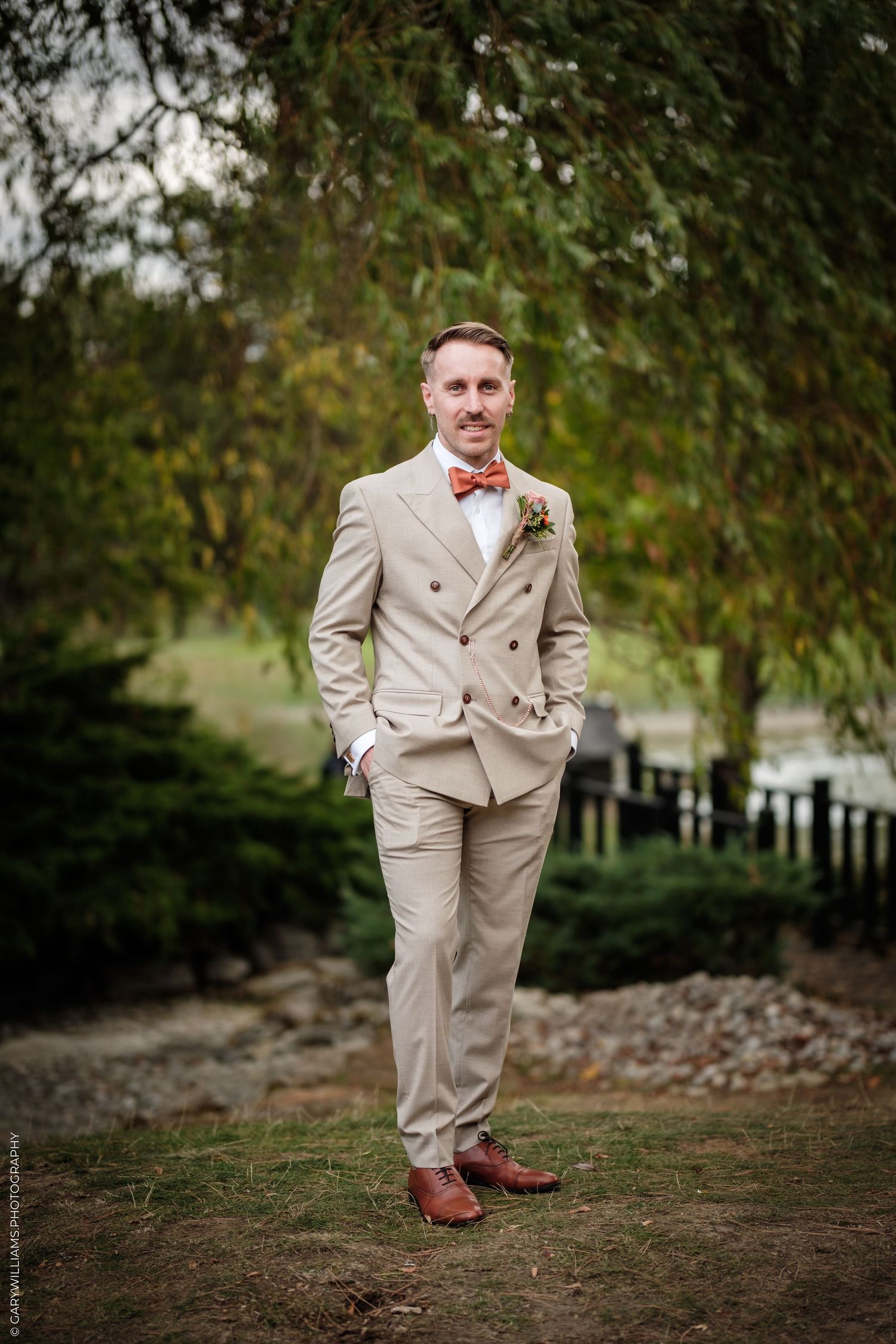 Groom in beige double-breasted suit with rust bow tie and boutonniere stands outdoors under willow tree.