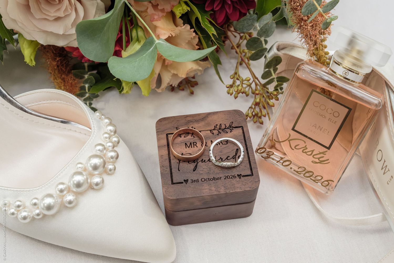 Wedding flat lay with pearl bridal heels, wooden ring box with gold bands, floral bouquet, and personalized perfume bottle.