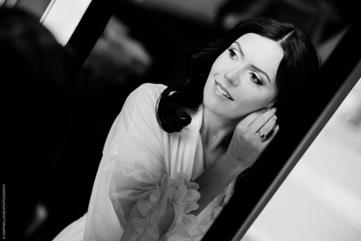 Black and white photo of a smiling dark-haired woman in a white robe, touching her face elegantly.
