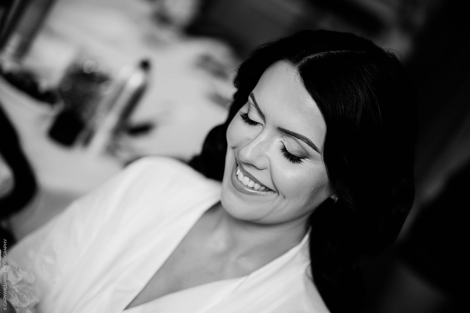Black and white photo of a smiling bride in a white robe, with elegant makeup and dark hair, captured during wedding preparations.
