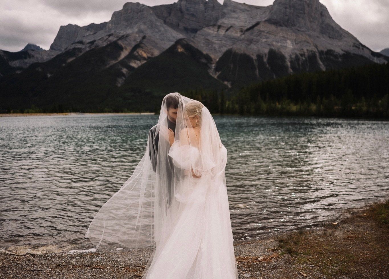Couple kissing under the bride's long veil on the shore of the Rundle Forebay in Canmore, Alberta