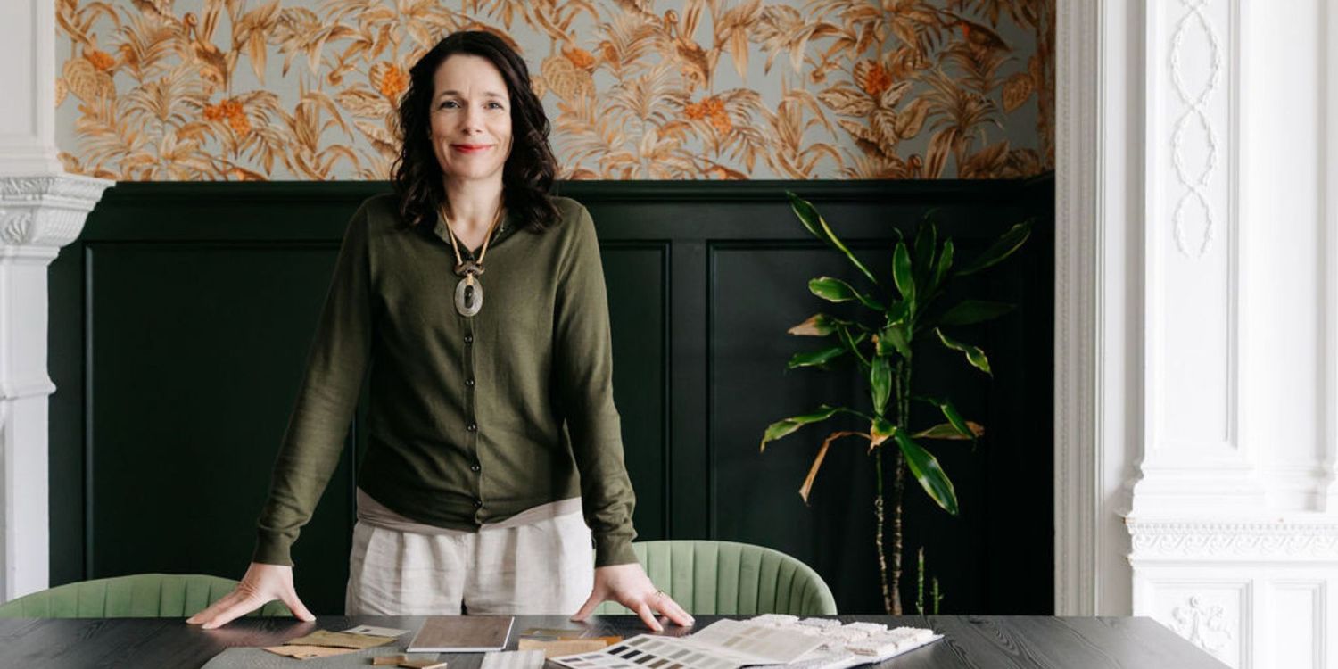 A woman in a green top stands confidently at a table in an elegantly decorated room with dark wainscoting and floral wallpaper.