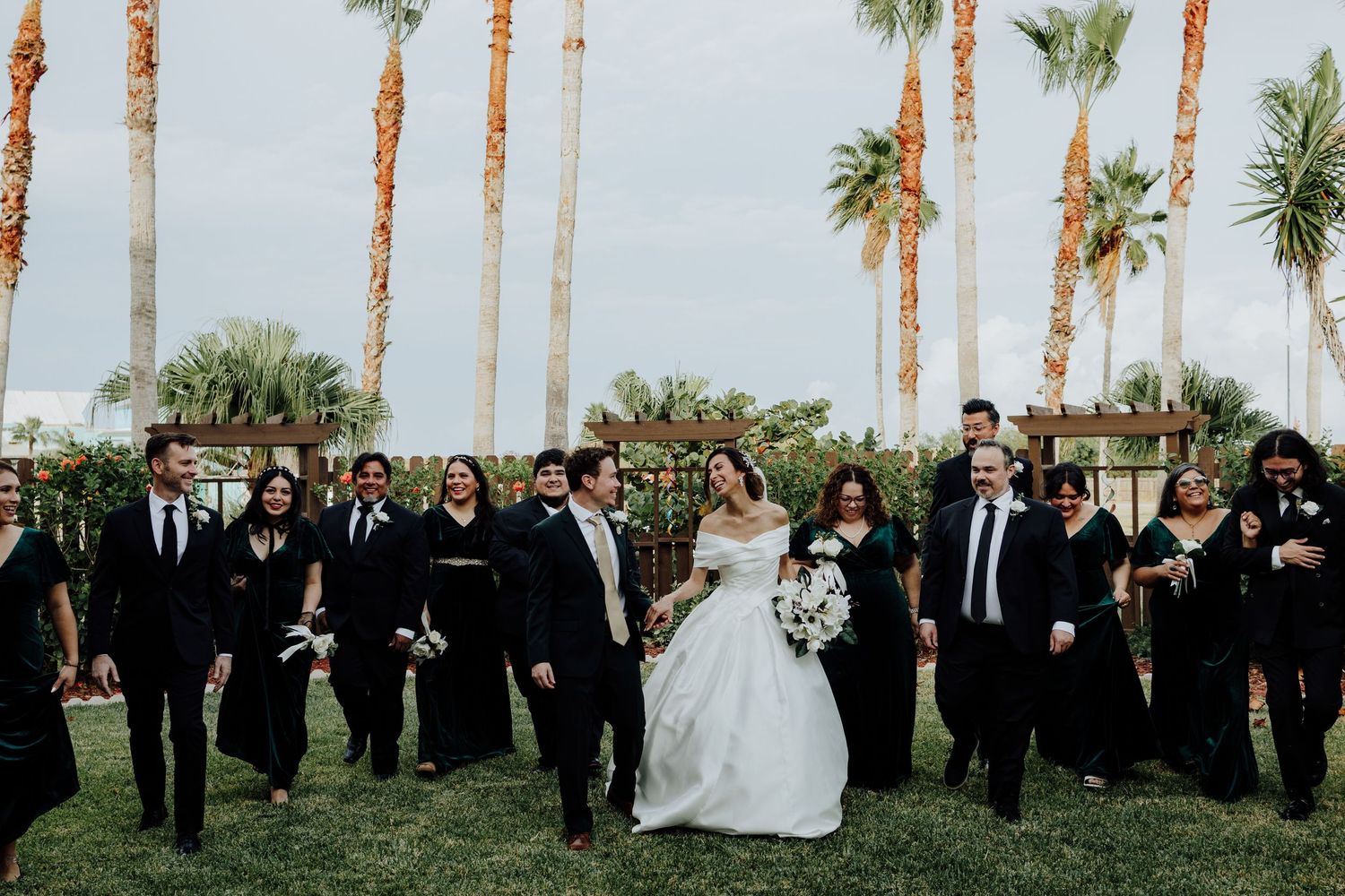 Wedding party in black attire walking with bride and groom near palm trees.