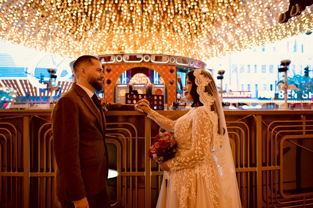 Bride in a lace gown and veil holding a bouquet stands facing a groom in a brown suit beneath a canopy of twinkling string lights.