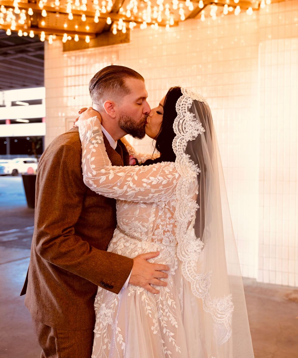 Bride in a lace veil and embroidered gown shares a romantic kiss with her bearded groom in a brown suit under warm string lights.