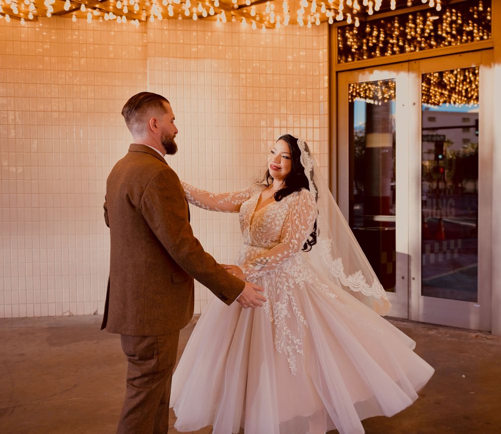A bride in a lace gown and veil twirls while dancing with her groom in a brown suit beneath warm string lights.
