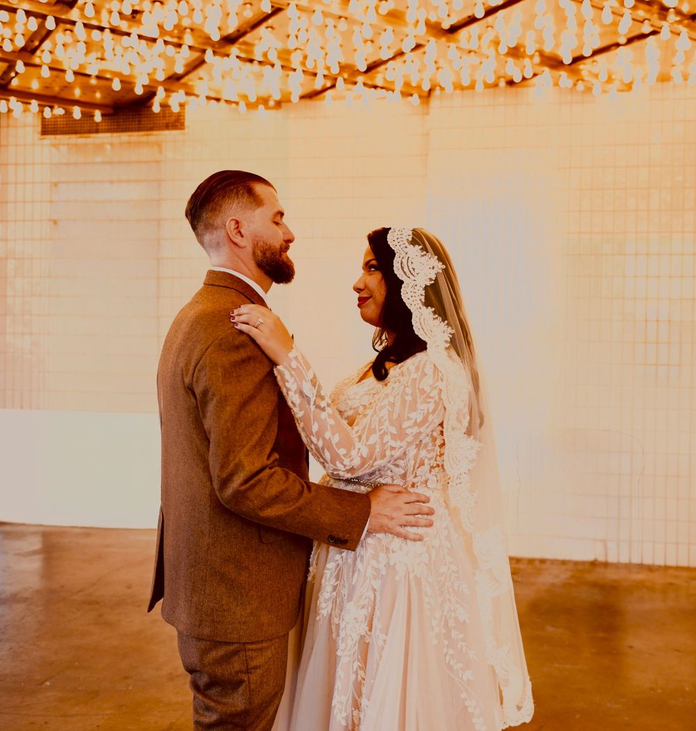 Bride in a lace gown and cathedral veil embraces a groom in a brown tweed suit under glowing string lights at a wedding.