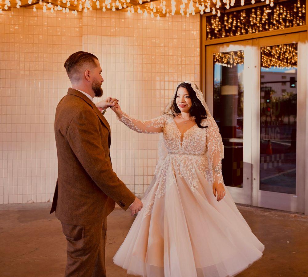 Bride in a blush lace wedding gown dances with a groom in a brown suit under warm string lights at a glass-door venue.