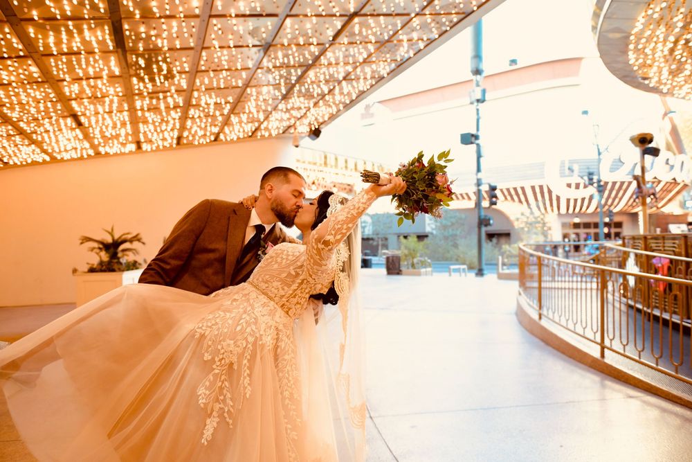Bride in a lace wedding gown and groom in a brown suit share a dipped kiss under a canopy of twinkling lights at an outdoor Las Vegas venue.