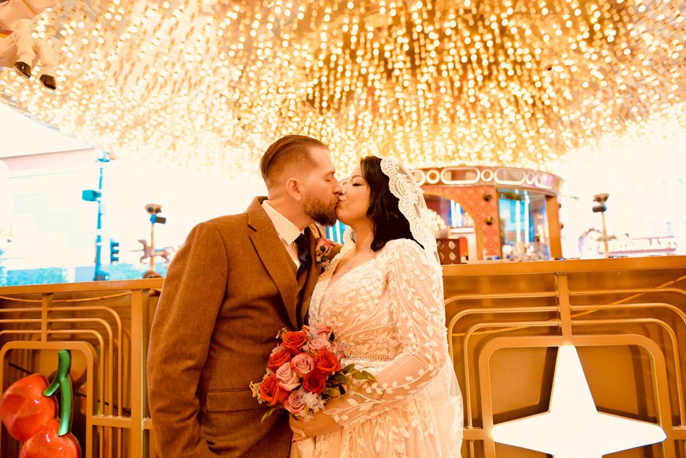 Bride and groom share a kiss under a glittering chandelier ceiling, she in a lace gown holding a red and pink bouquet, he in a brown suit.