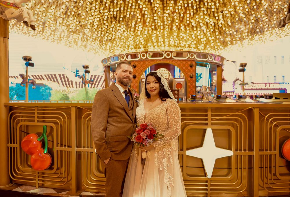 Bride in a lace gown with veil and groom in a brown suit hold a red bouquet at a retro Las Vegas wedding chapel with glowing gold lights.