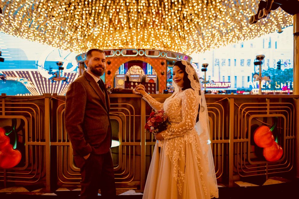 Bride in a lace wedding gown holding a bouquet stands beside a groom in a brown suit under glowing string lights at a retro neon-lit venue.
