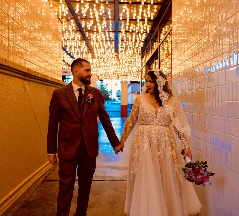 Bride in a lace gown and veil holds hands with her groom in a brown suit while walking under string lights in a tiled passage.