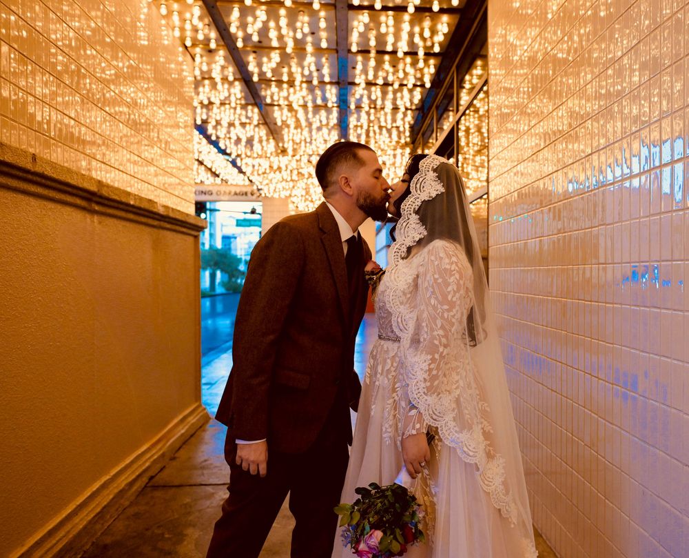 Bride in a lace gown and veil kisses her groom in a brown suit in a tiled passage glowing with string lights on their wedding day.