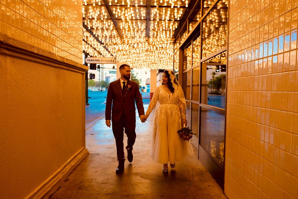 Newlywed couple holding hands and a bouquet stroll down a warmly lit tiled passage beneath twinkling canopy lights.