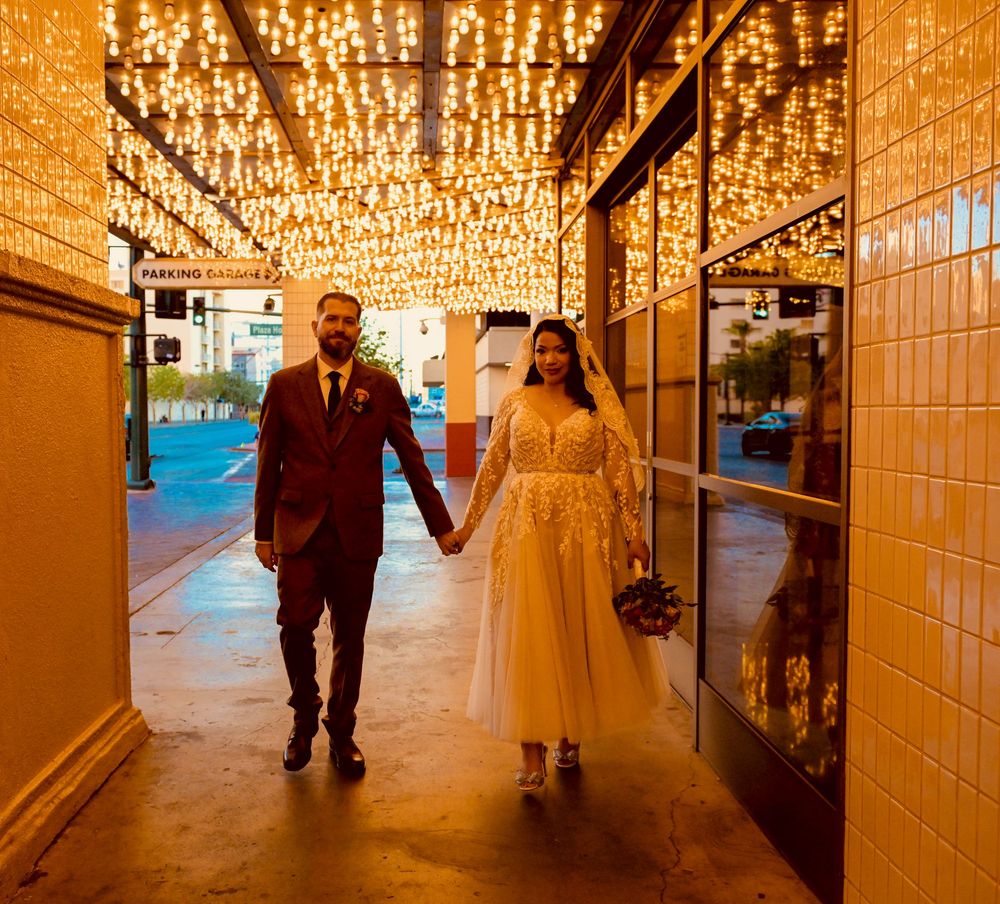 Bride in a lace wedding gown and groom in a burgundy suit hold hands under a glowing marquee of string lights on a Las Vegas street.