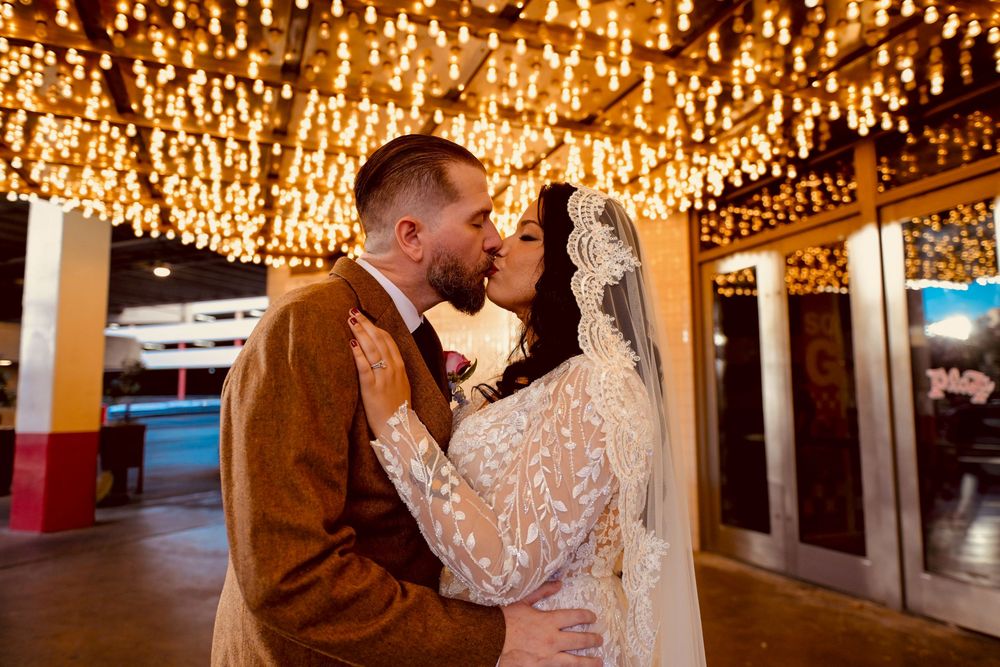 Bride in a lace veil and groom in a brown suit share a kiss under glowing string lights at a Las Vegas-style wedding.