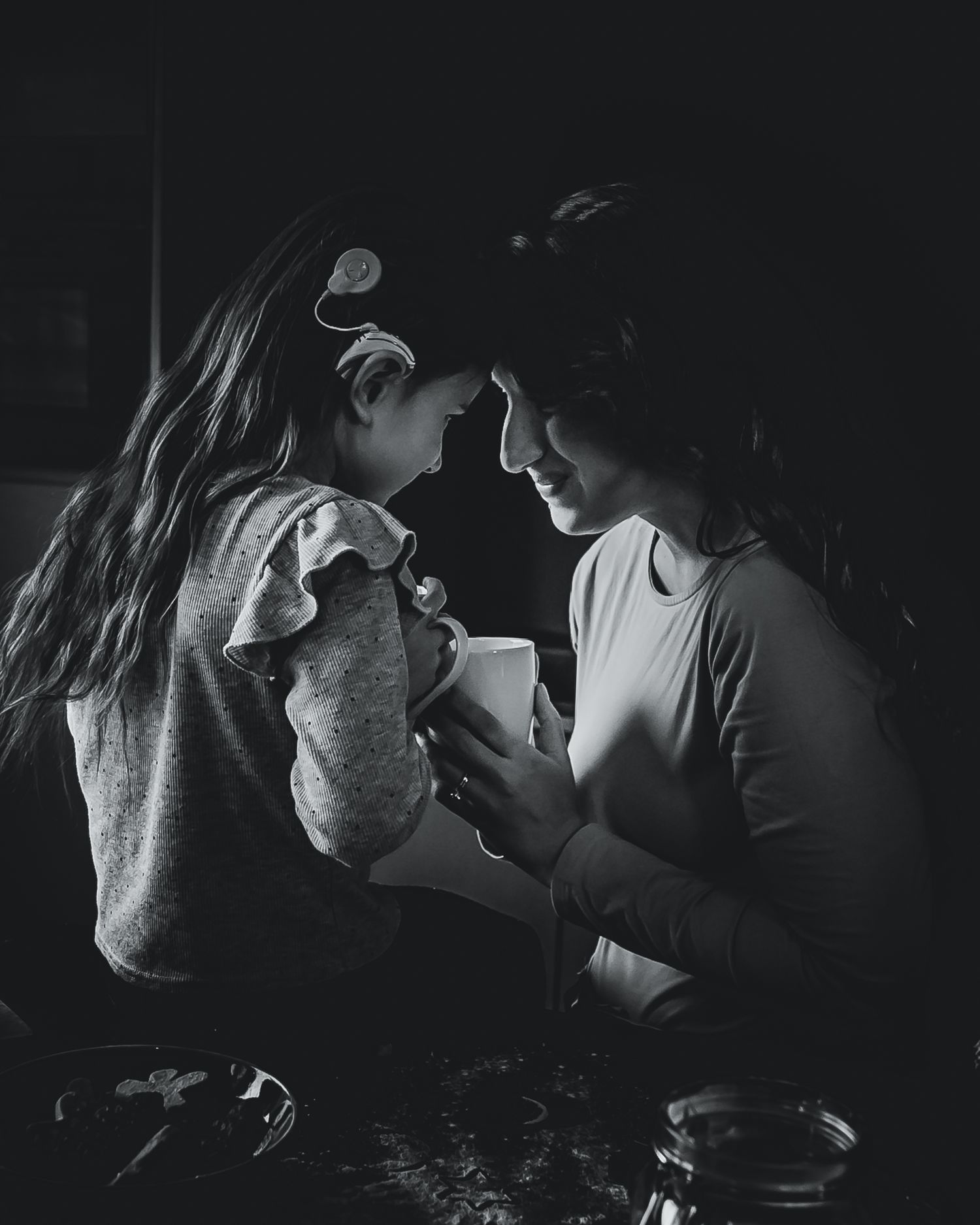Moody photo of a young girl with a hearing device touching foreheads with a smiling mother as they drink hot chocolate together in Utrecht.