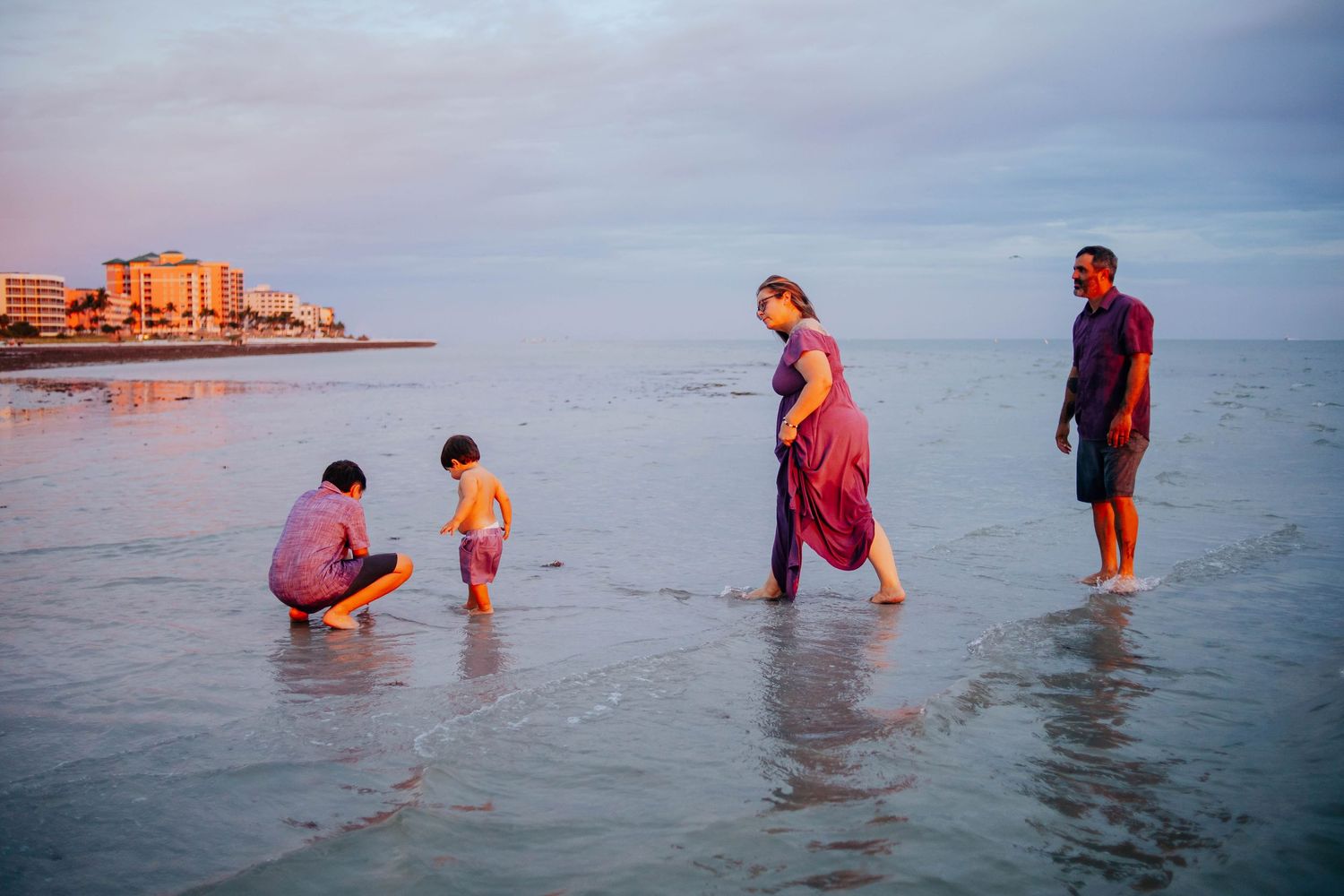 A family wades in shallow ocean water at sunset, with two children crouching and two adults standing near a distant beachfront skyline.