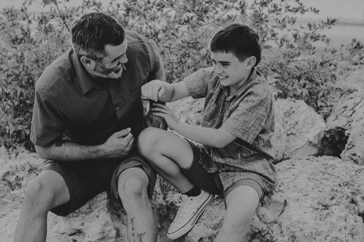 Black and white photo of a smiling man and a young boy sitting together on large rocks, laughing while tying a shoe outdoors.