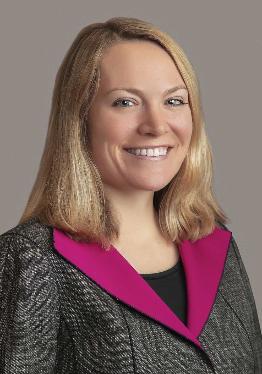 Smiling woman with shoulder-length blonde hair wears a gray tweed blazer with magenta lapels in a professional studio headshot.
