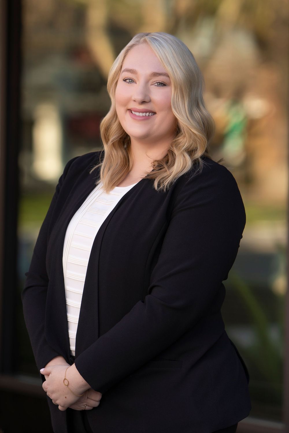 Smiling woman with long wavy blonde hair wearing a black blazer over a white top, posing outdoors in a professional headshot.