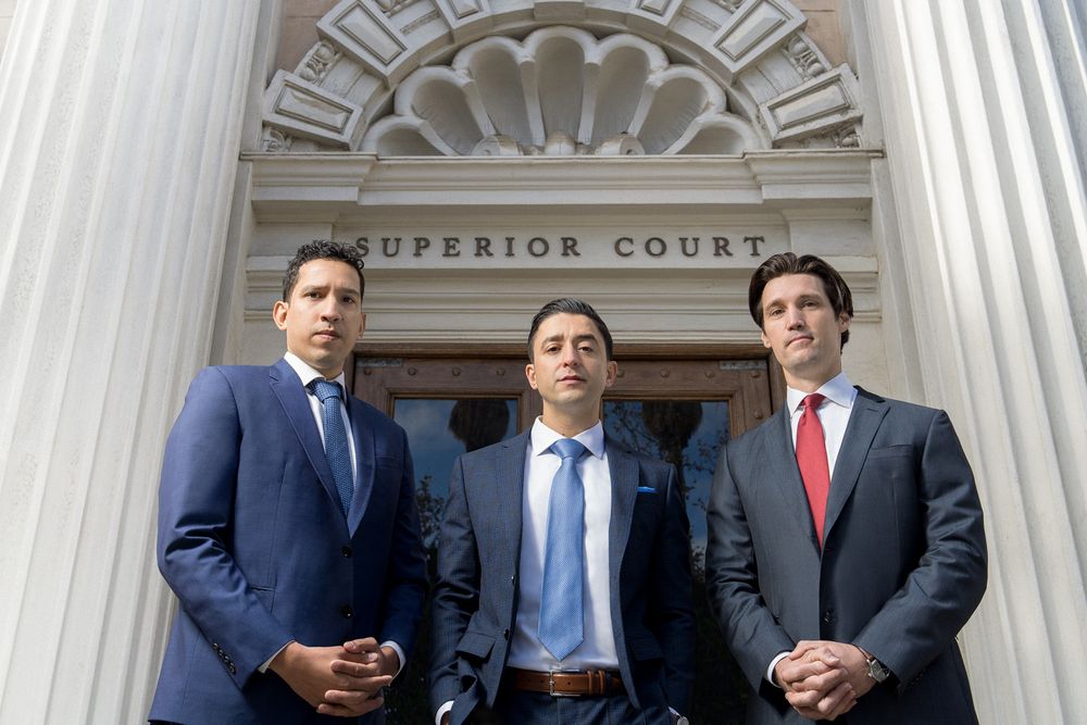 Three men in dark business suits and ties stand together in front of the Superior Court building entrance between tall stone columns.