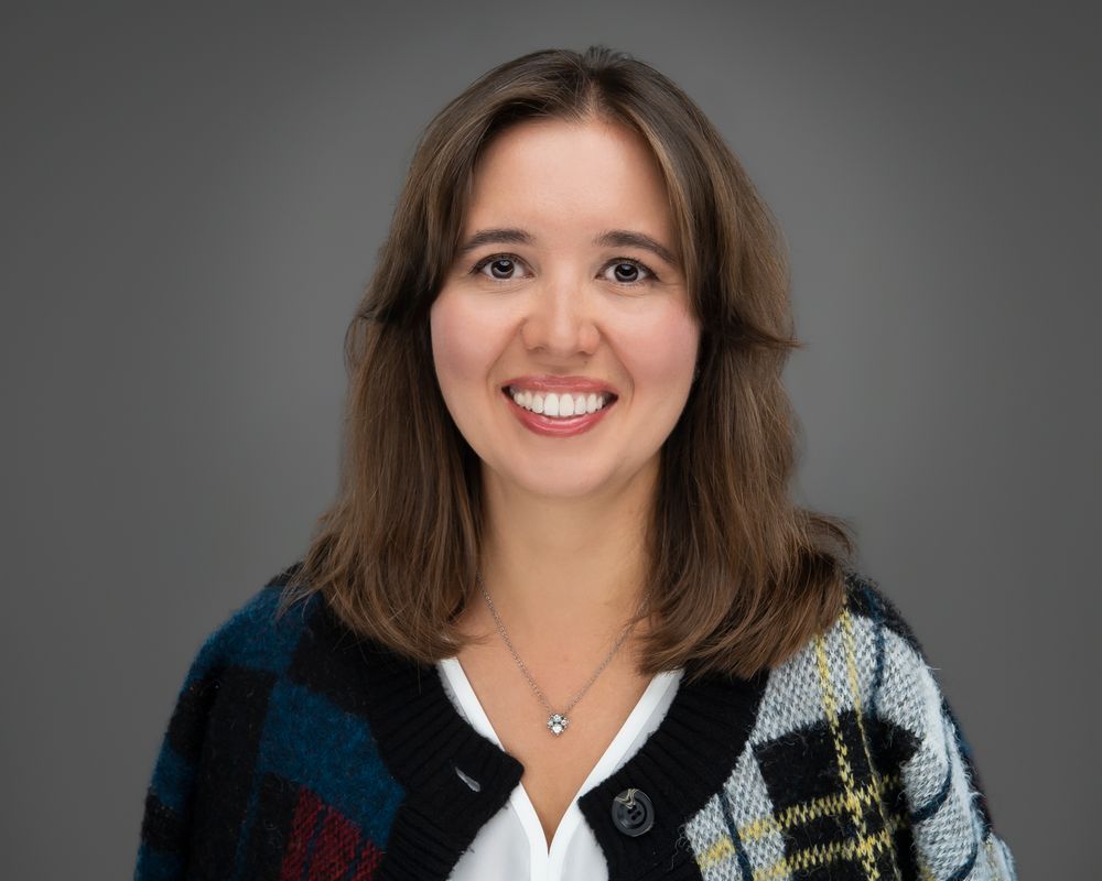Smiling woman with shoulder-length brown hair wearing a colorful patterned cardigan over a white top and a delicate pendant necklace.