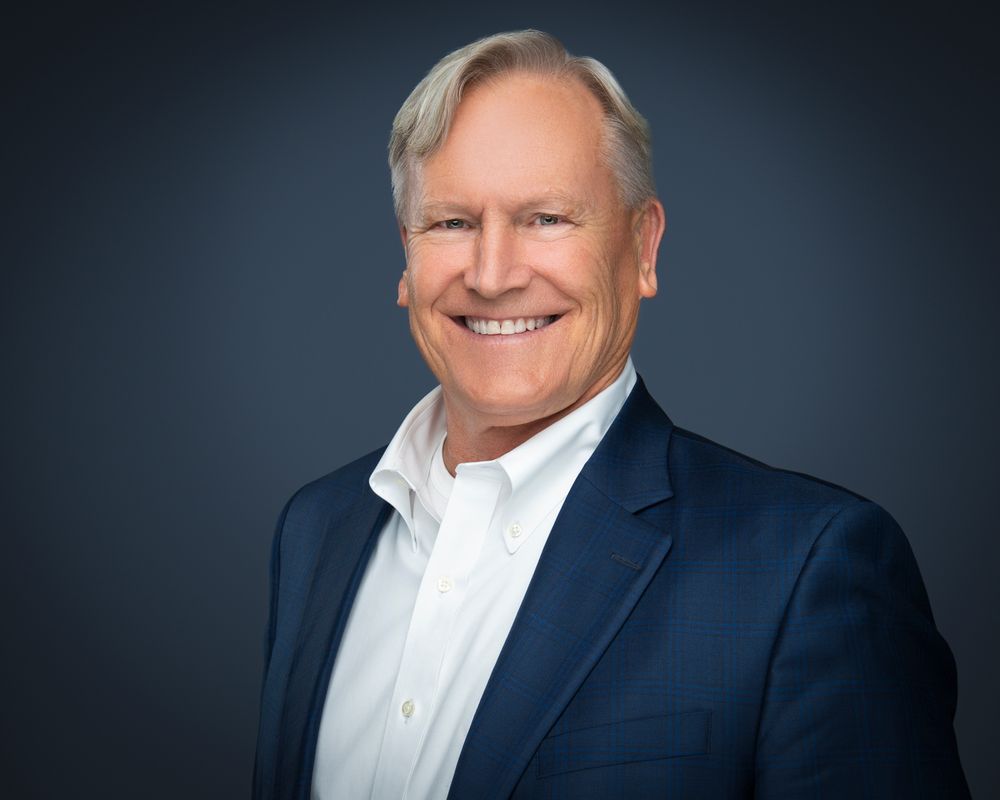 Smiling gray-haired man in a navy blazer and white open-collar shirt posing for a professional headshot against a dark background.