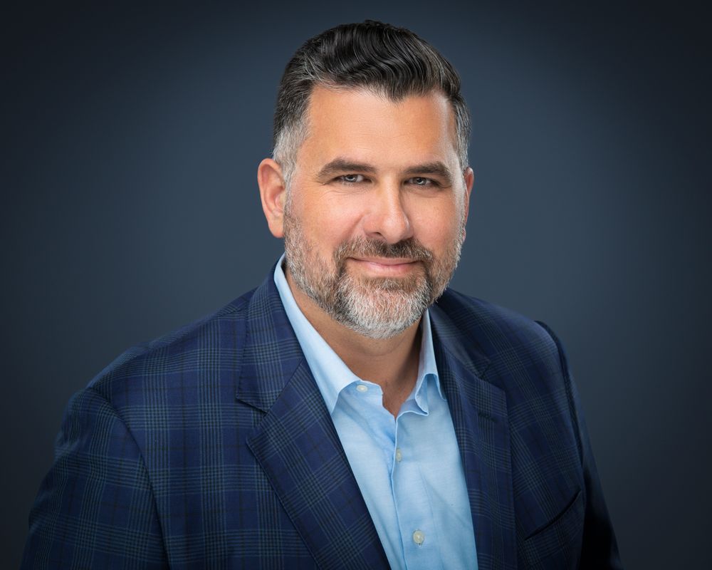 Professional headshot of a smiling man with short dark hair and a graying beard wearing a navy plaid blazer and light blue collared shirt.