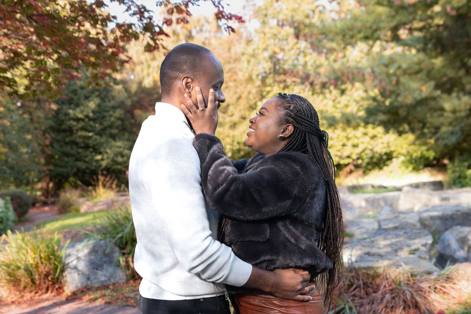 A couple shares a tender moment outdoors, embracing among autumn trees in a lush garden setting with engagement ring after recent proposal