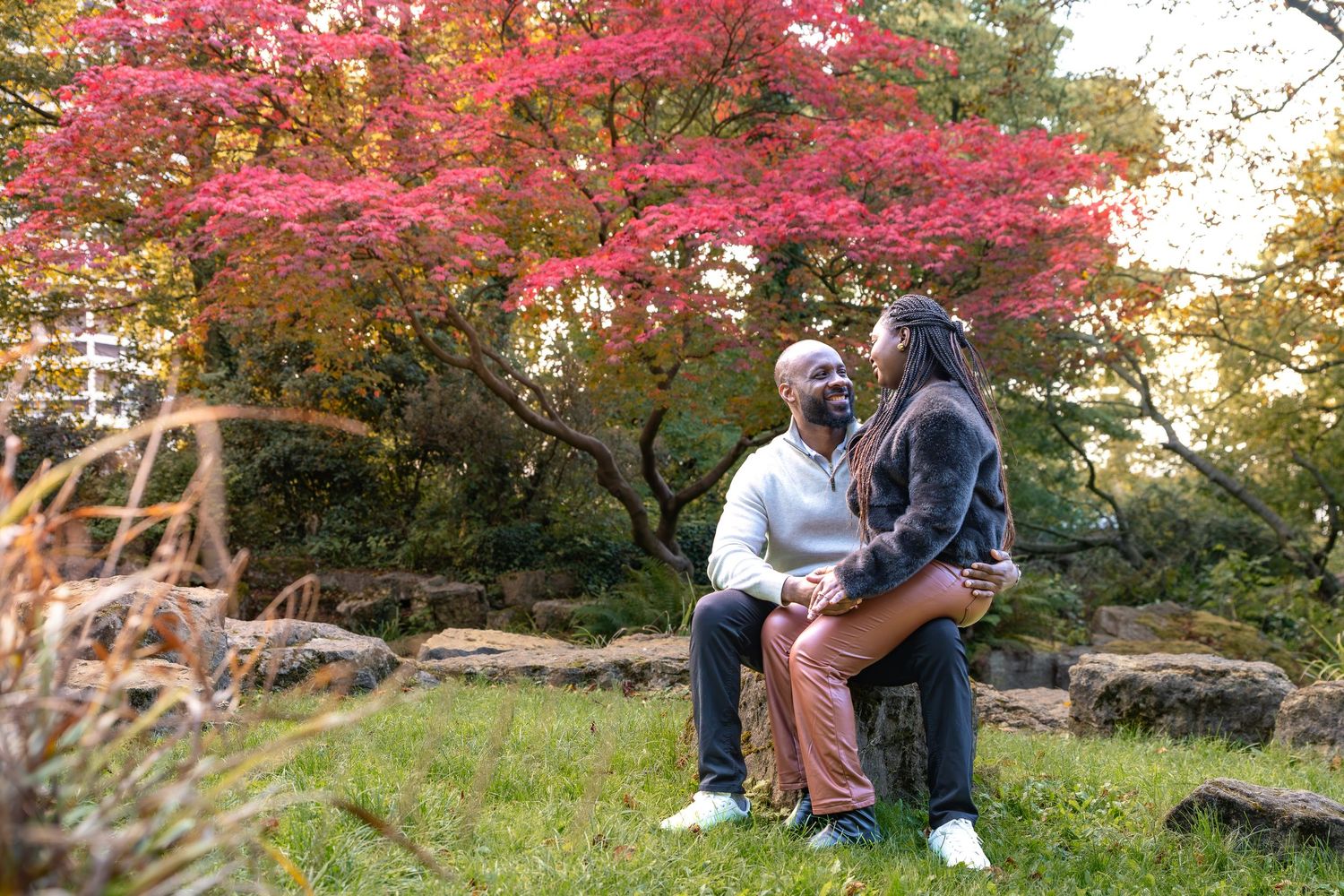 Couple sitting together in a lush autumn garden with vibrant red Japanese maple trees in the background showing engagement ring Rotherham