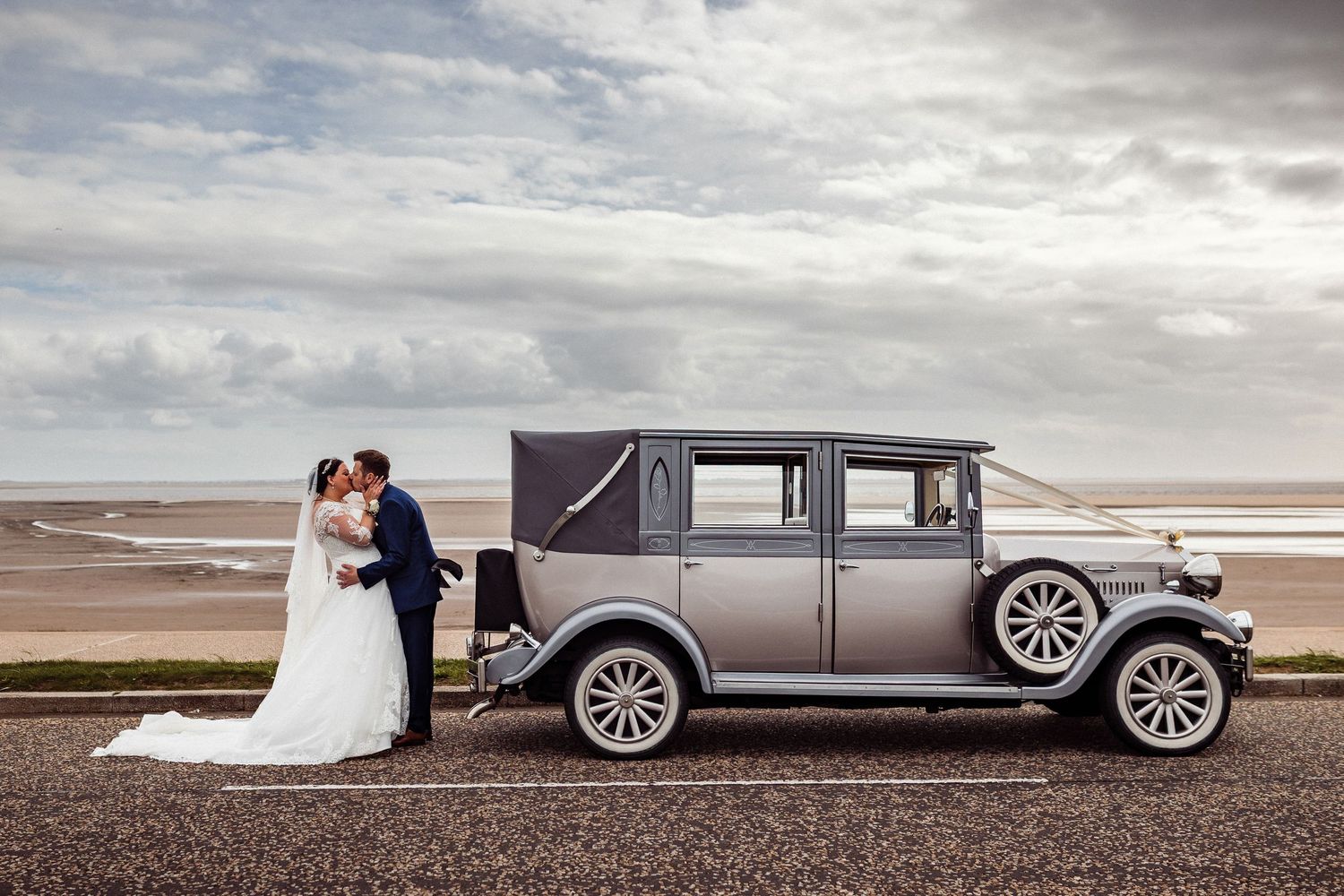 Bride and groom kissing beside a vintage gray wedding car on a coastal road with sandy beach backdrop.