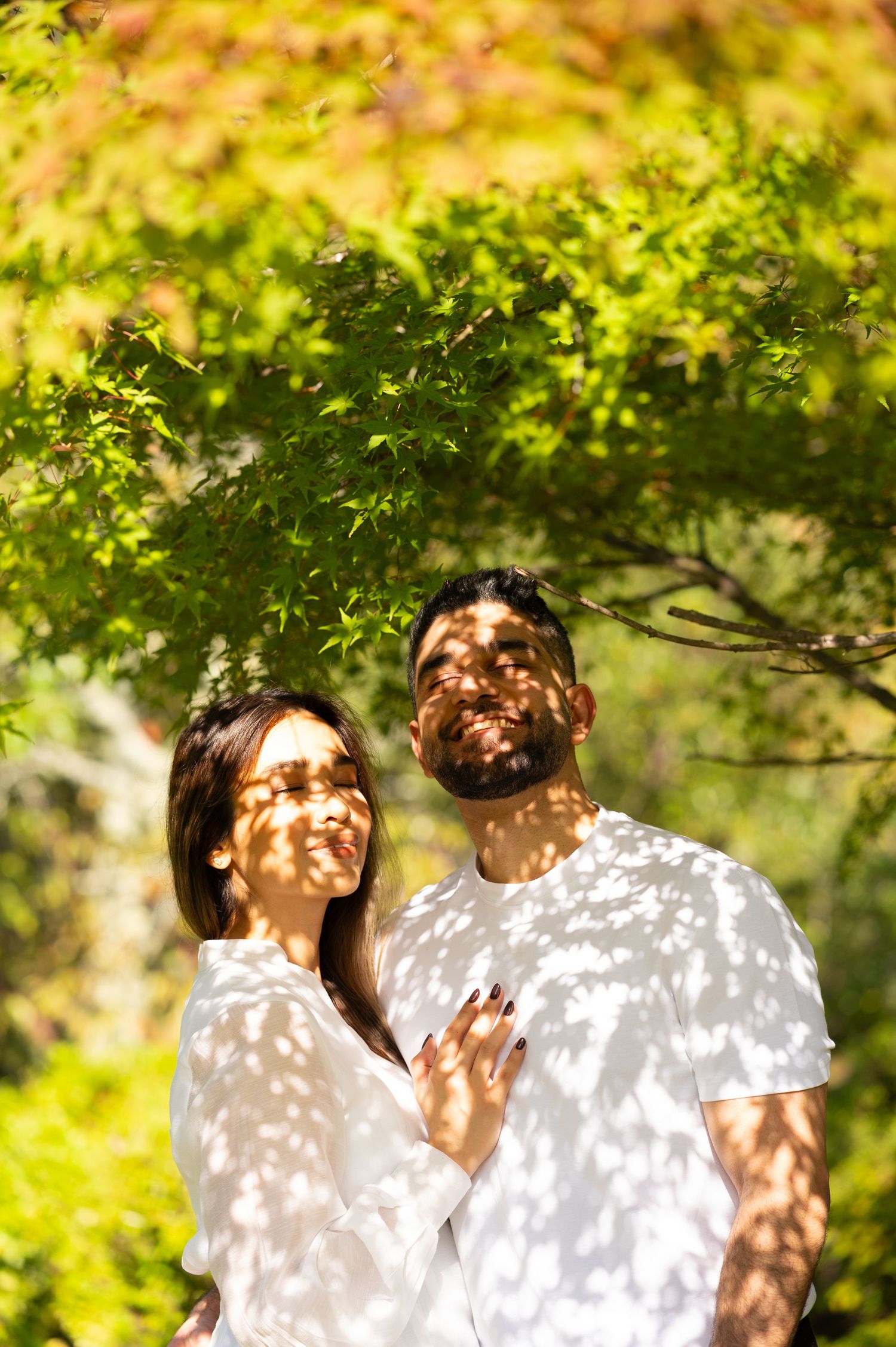 Smiling couple in white outfits embrace under green and golden autumn leaves as dappled sunlight casts leaf shadows on their faces.